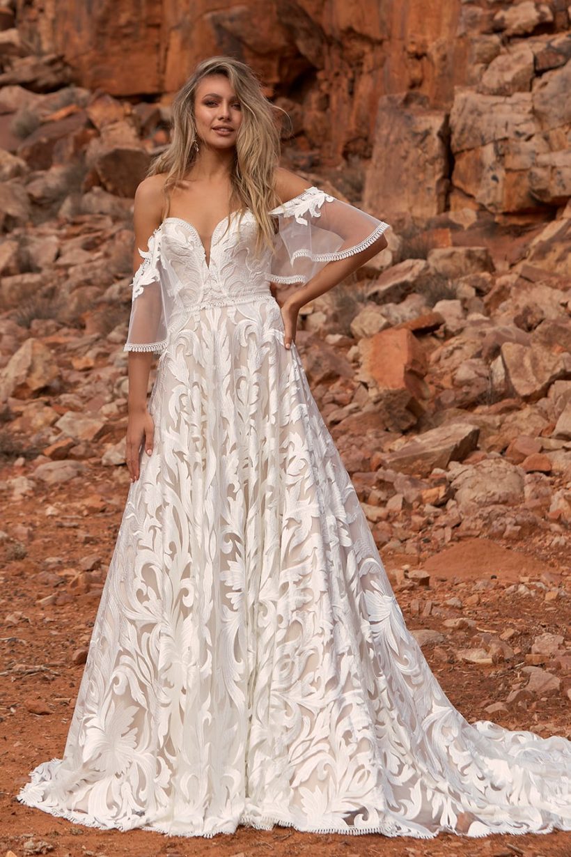 Különleges hímzett csipkével díszített A vonalú menyasszonyi ruha mély - tüllbetéttel kombinált -szív alakú kivágással. Érdekessége a bő, levehető csipkeujj, mely bohém külsőt kölcsönöz a modellnek. Egy igazán rendkívüli darab az Evie Young esküvői ruha kollekcióból! Style: Wyatt. Elölről fotózva
