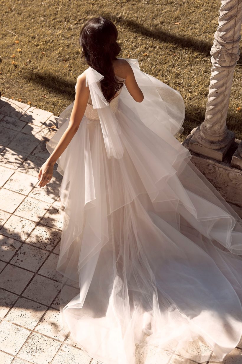 Romantikus, fodros tüll menyasszonyi ruha modern hercegnőknek, flitterekkel díszített csipkebodyval és félvállas tüll kiegészítővel a Madi Lane 2021 évi menyasszonyi ruha kollekcióból. Style: Bella. Hátulról fotózva