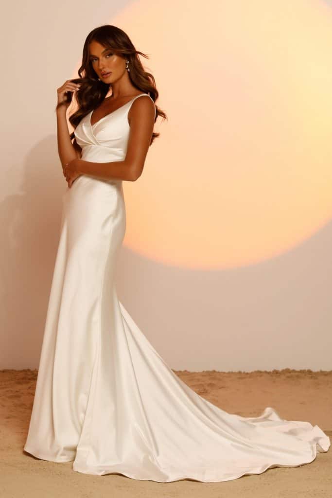 A vonalu, vállpántos V nyakkivágású lágy selyem menyasszonyi ruha mutatós uszállyal és széles övvel a derekán. Style: Jaxon a Madi Lane 2023 évi "Oasis" kollekcióból oldalról
