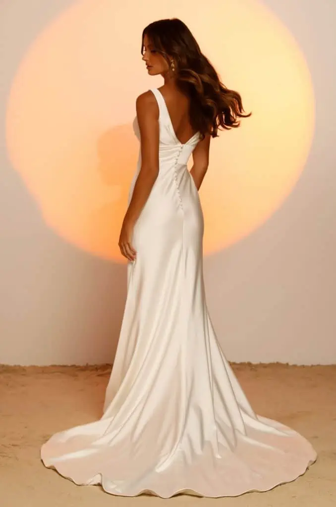 A vonalu, vállpántos V nyakkivágású lágy selyem menyasszonyi ruha mutatós uszállyal és széles övvel a derekán. Style: Jaxon a Madi Lane 2023 évi "Oasis" kollekcióból Hátulról