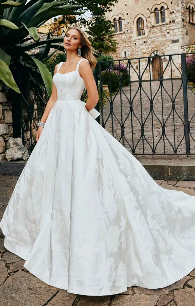 Sima, egyszerű esküvői ruha brokát anyagból. Bodyja szolíd kivágású, széles vállpántú, a ruha hátuját pedig masni díszíti. Style: Veni Infantino "52075"